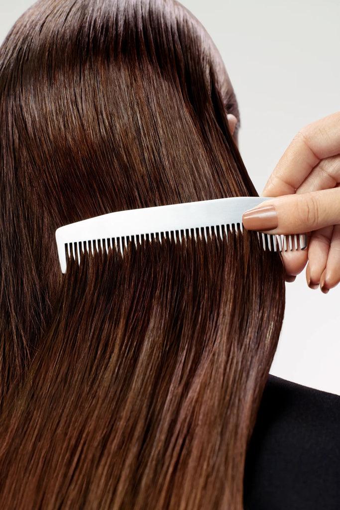 Person combing their long brown hair with a white comb on a neutral background